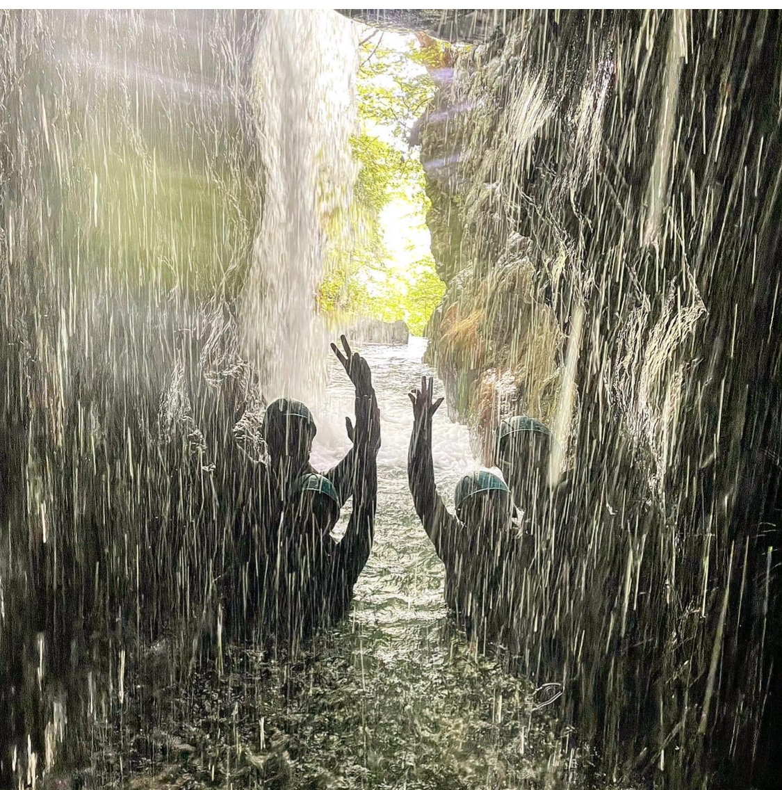 A group of people standing in mountain stream with their arms raised with a waterfall in from of them.