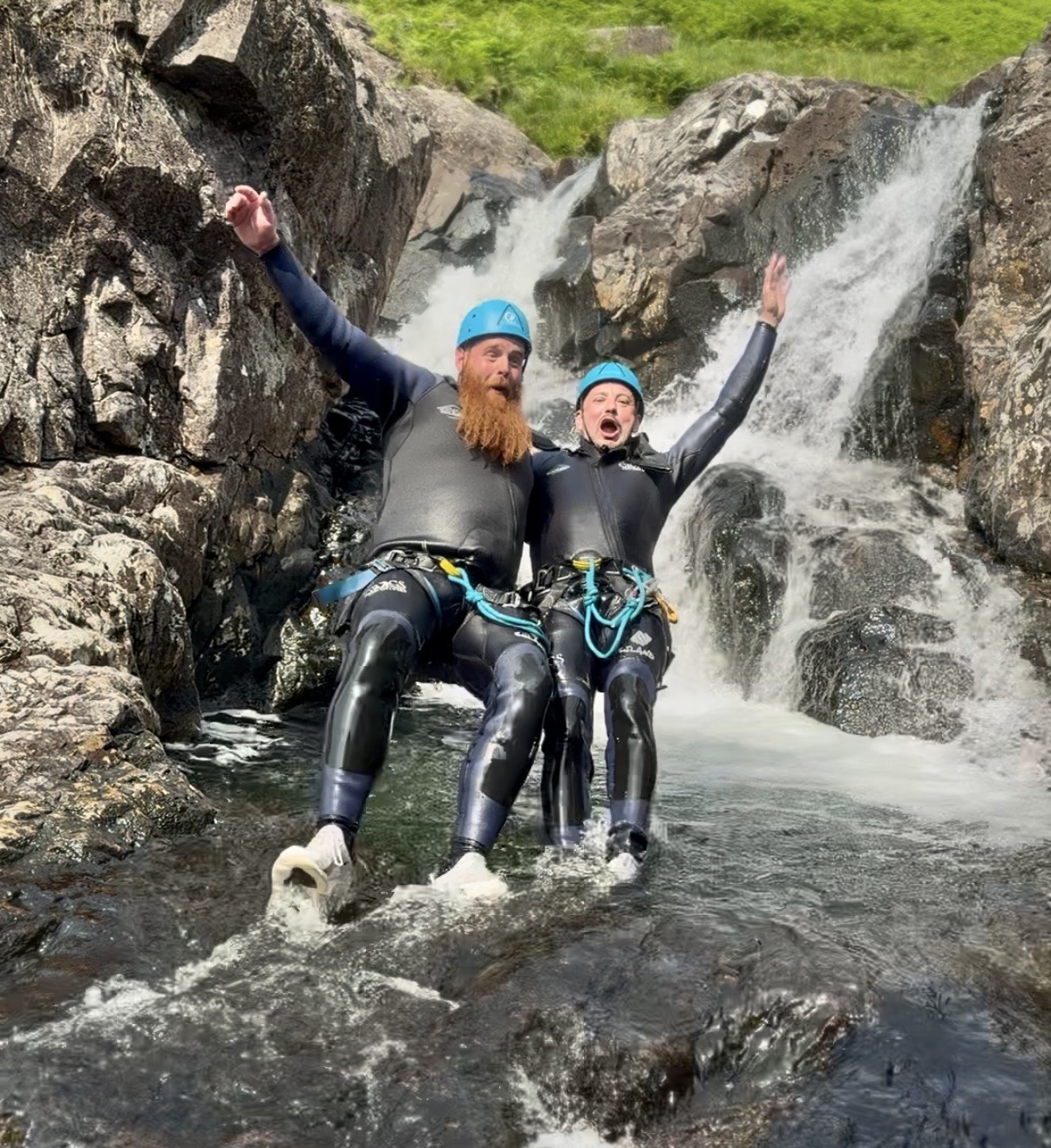 two male adult friends fall backwards into a mountain stream with one of their arms over each others shoulders