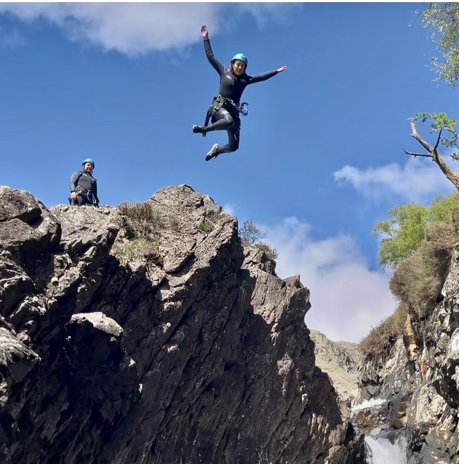 a woman jumping with her arms in the air off a high cliff jump in the lake district at esk ghyll while on an organised ghyll scrambling session